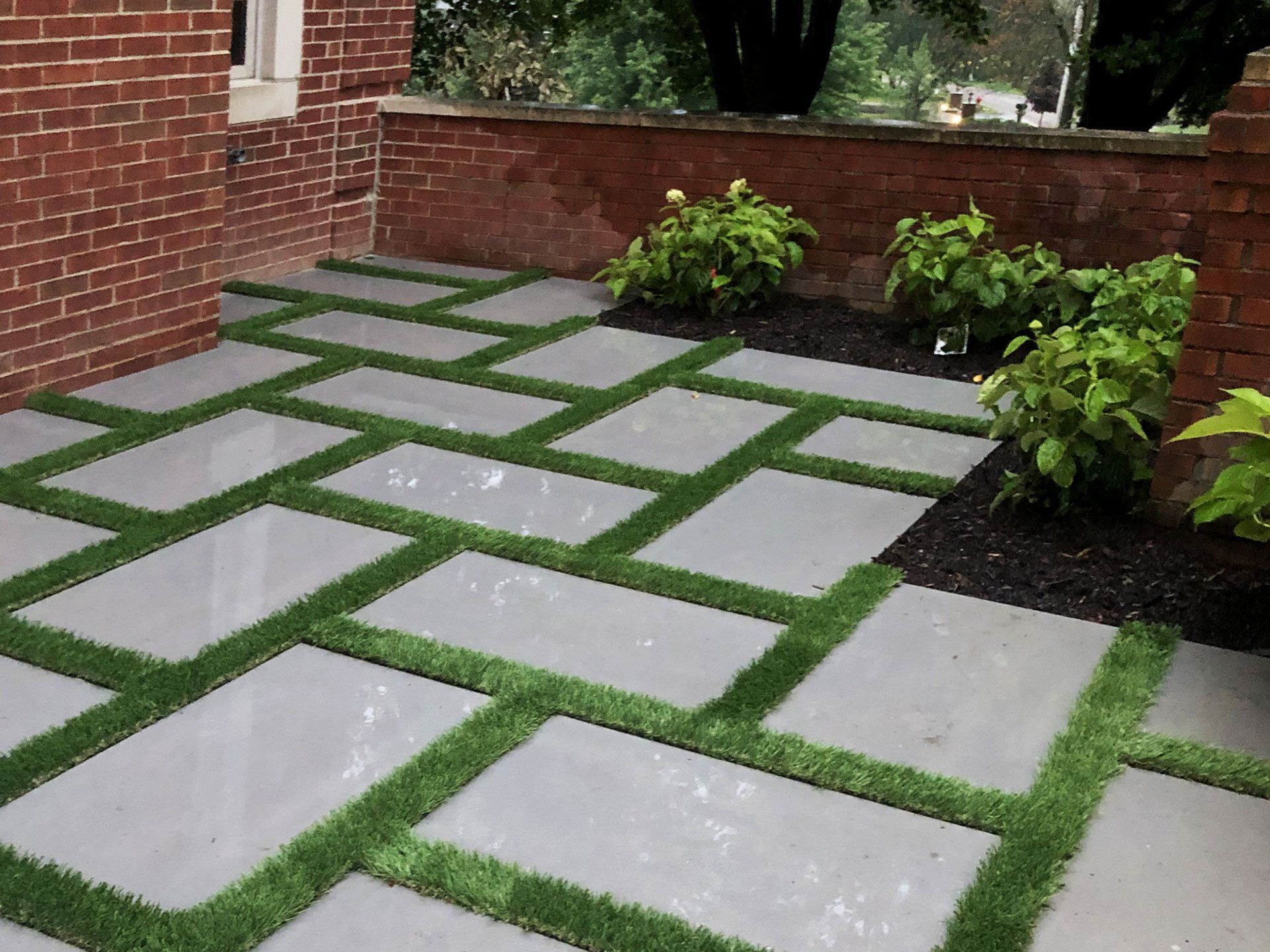 A patio with a brick wall and grass between the tiles