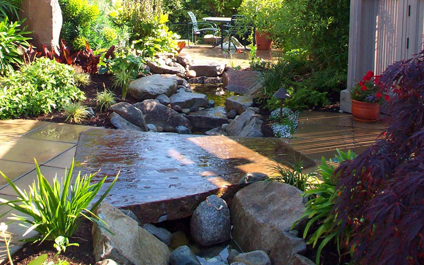 A garden with a stream running through it and a table and chairs in the background.