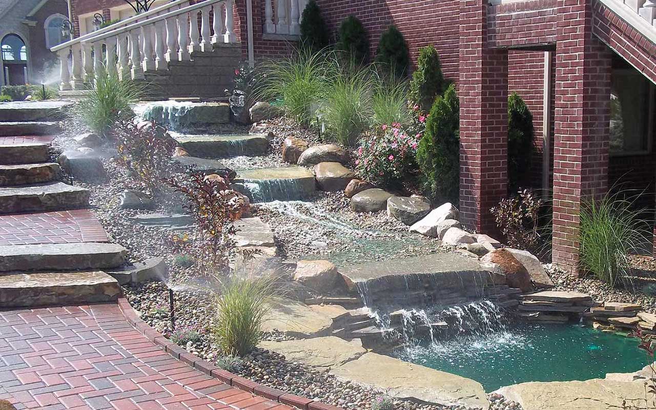 a waterfall is surrounded by rocks and stairs in front of a brick building .