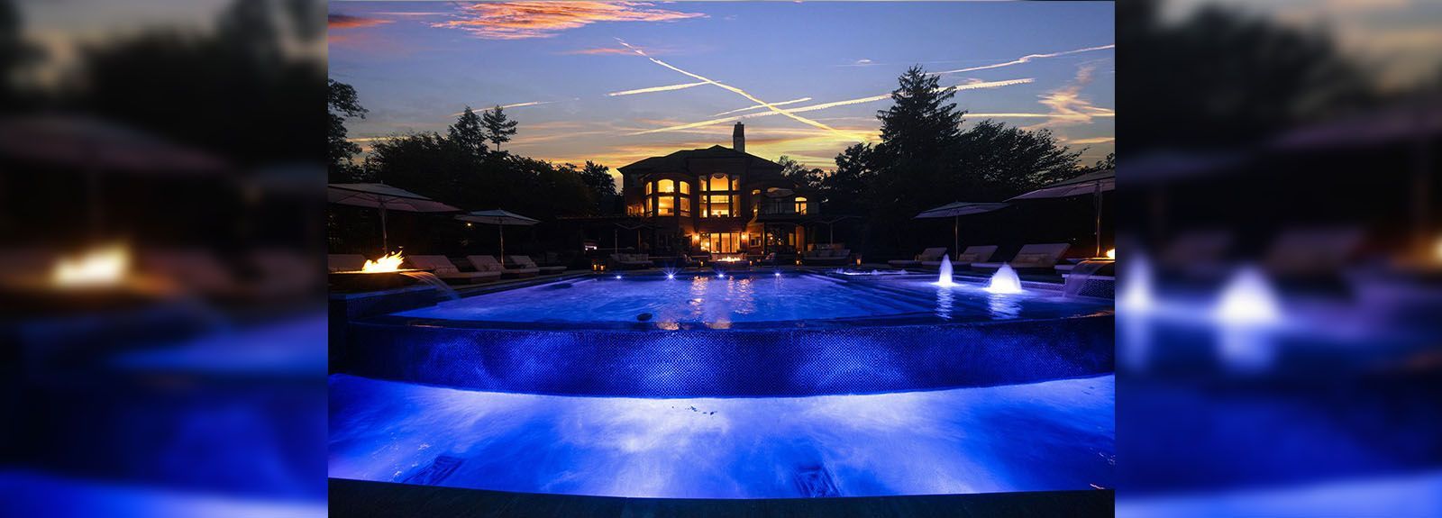 A large swimming pool is lit up at night with a house in the background.