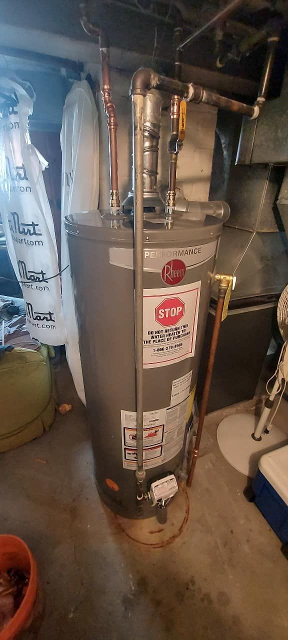 Basement water heater with silver tank, pipes, and red labels next to a laundry area.
