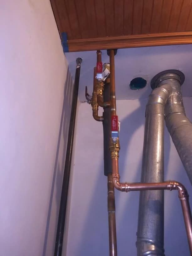 Exposed copper pipes and a valve beside a gray duct in a closet corner.