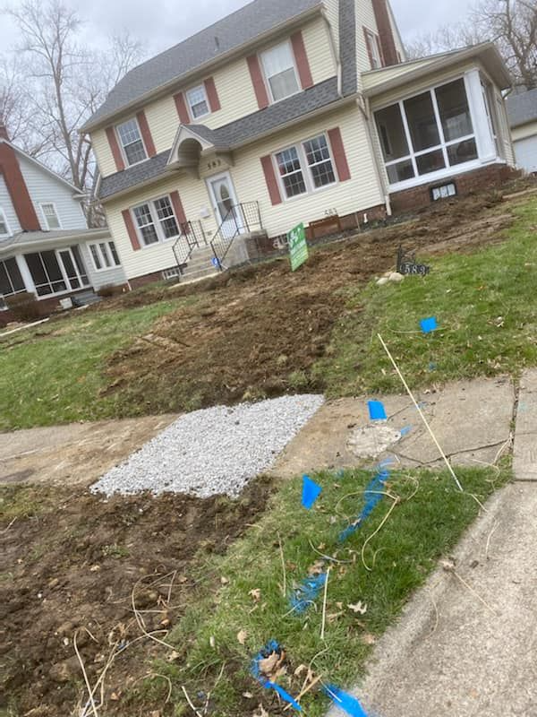 Residential yard with bare earth, gravel patch, and blue survey flags in front of a house
