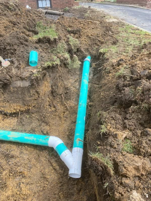 Exposed turquoise PVC pipes in a muddy trench beside a road, showing a Y-shaped junction.