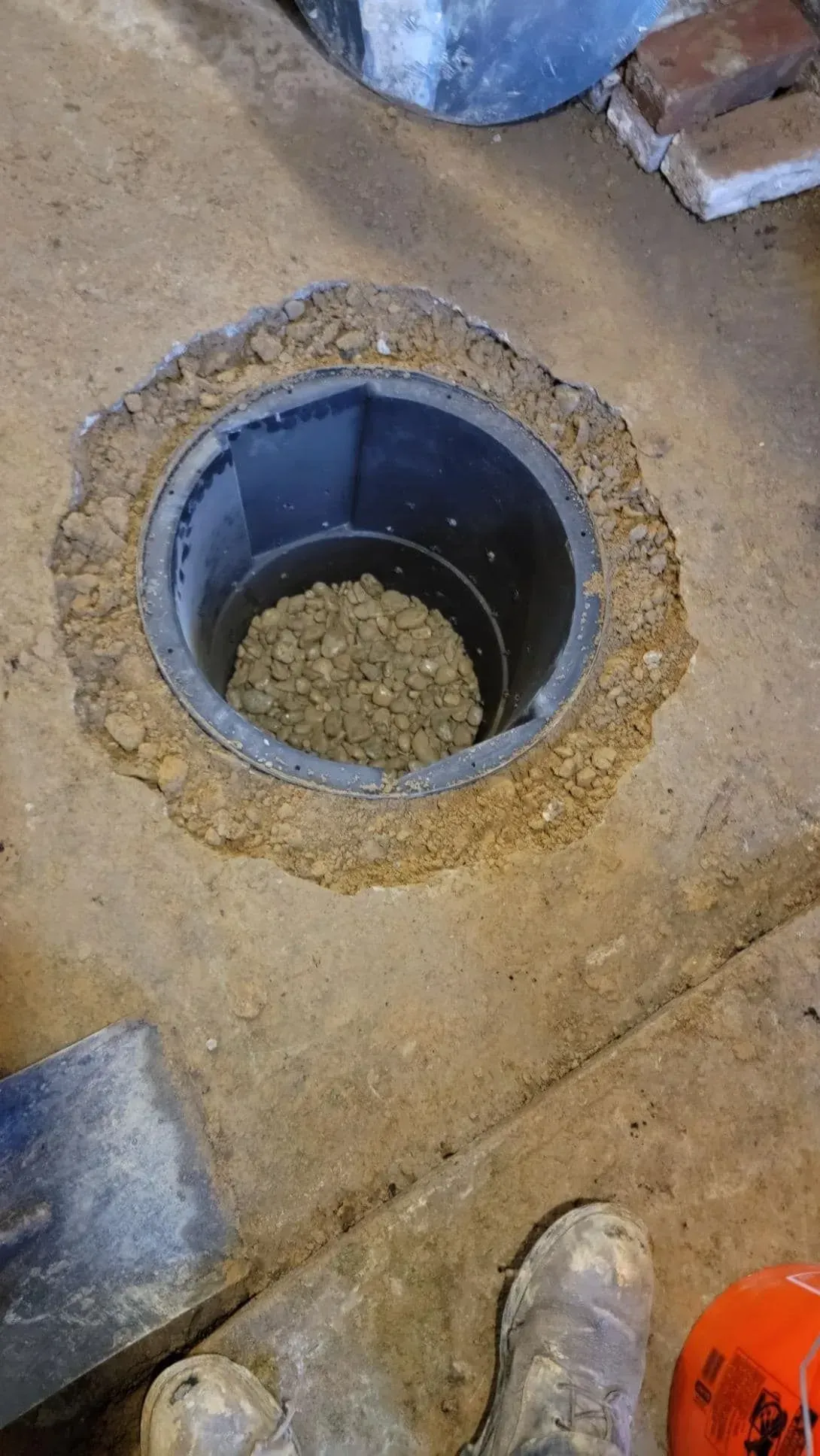 Open floor drain hole in a concrete floor with gravel inside, near a work boot and orange tool.