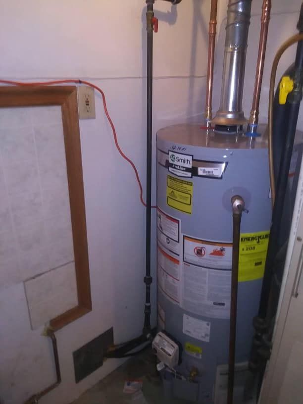Water heater in a narrow utility closet with exposed pipes and tiled walls.