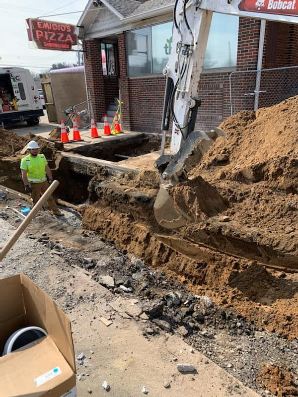 Construction crew repairing a collapsed sidewalk and excavation beside storefronts, with cones and a backhoe bucket visible
