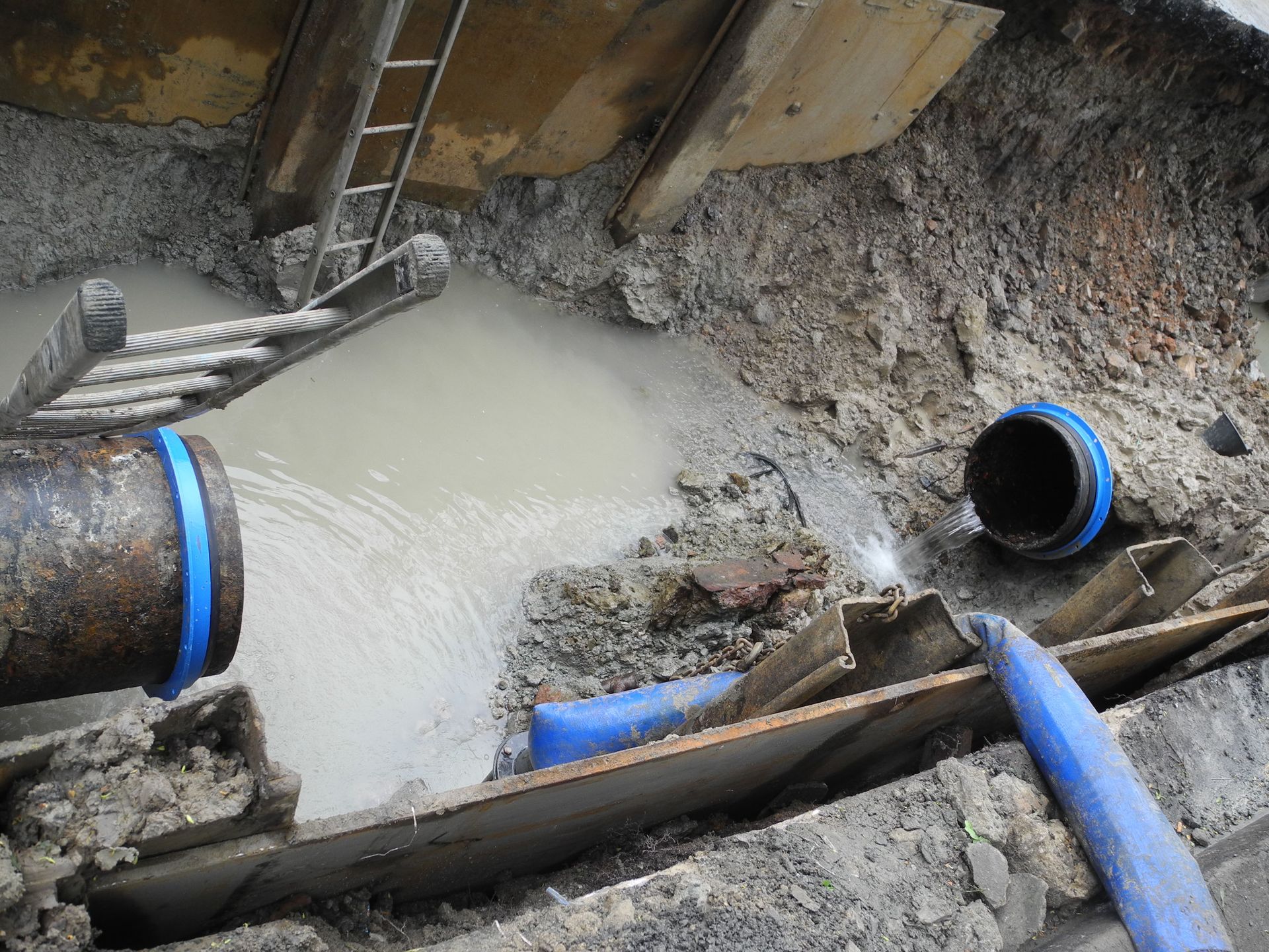 Flooded excavation site with muddy water, pipes, and a ladder against a dirt wall