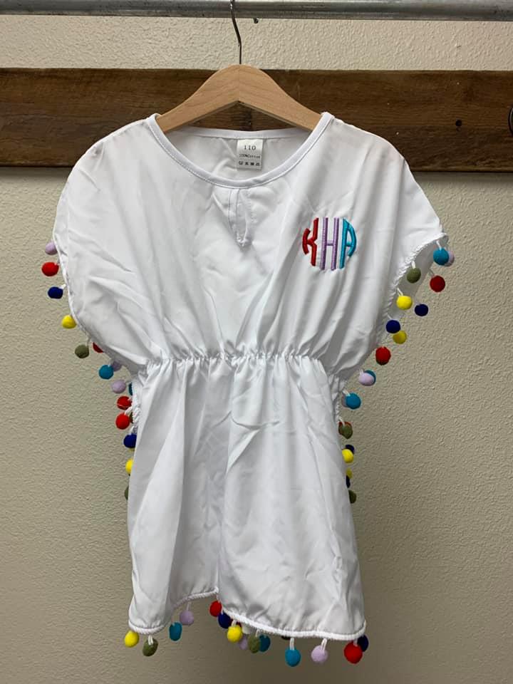 A white shirt with a monogram and colorful pom poms is hanging on a hanger.