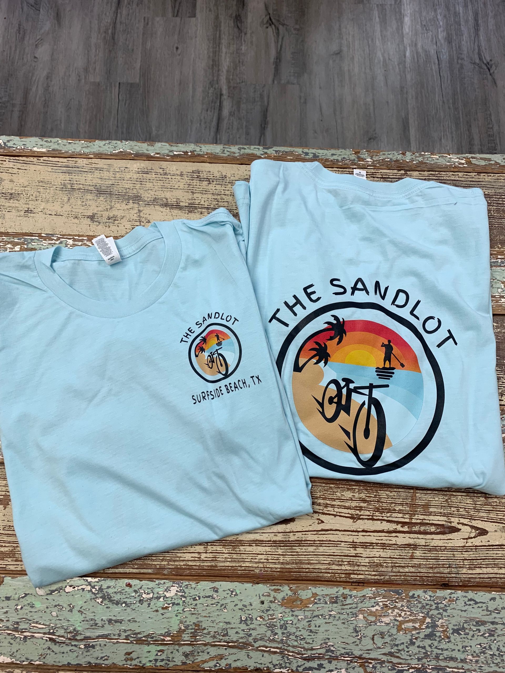 Two light blue t-shirts are sitting on a wooden table.