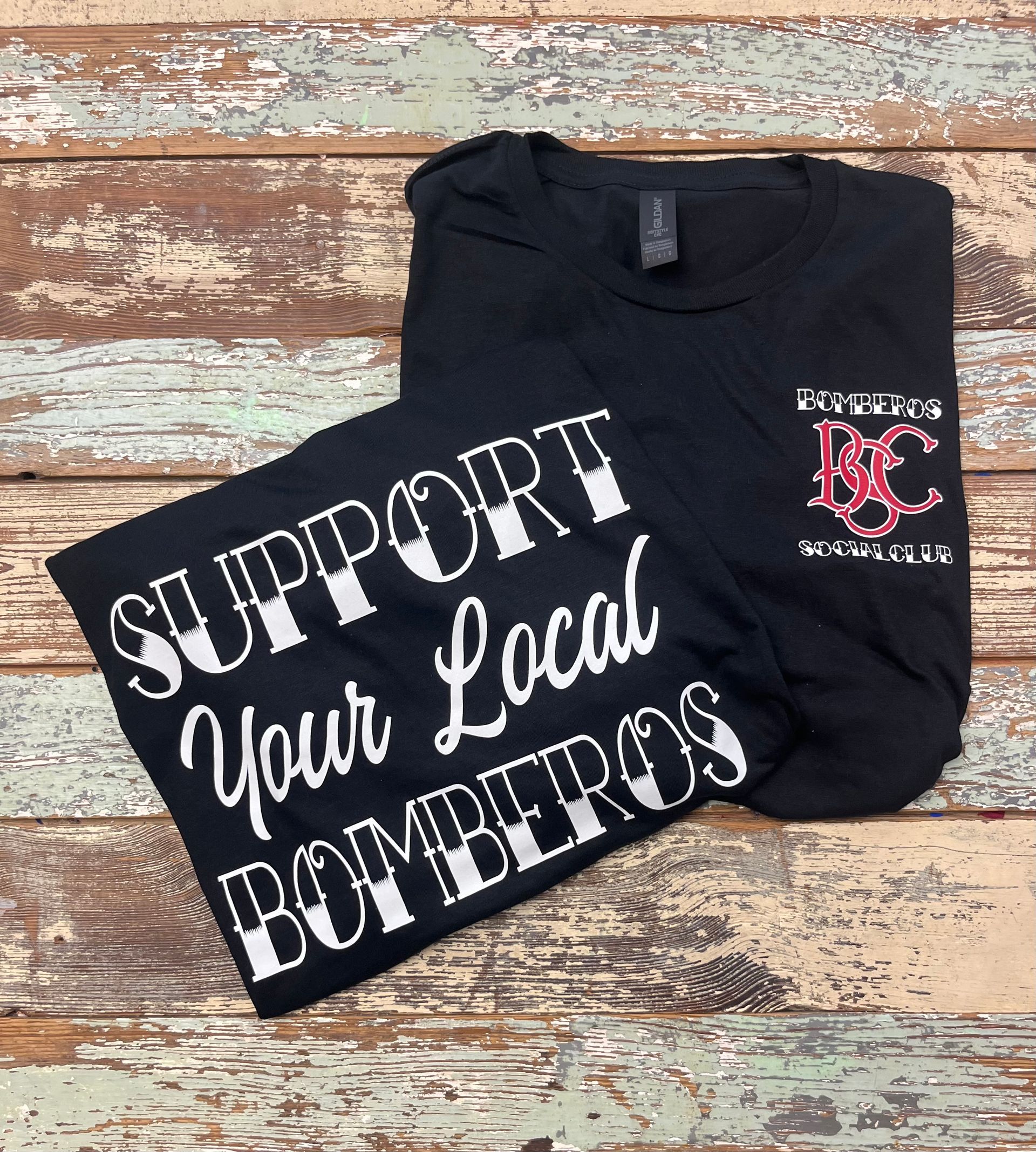 A black t-shirt that says `` support your local bomberos '' is sitting on a wooden table.