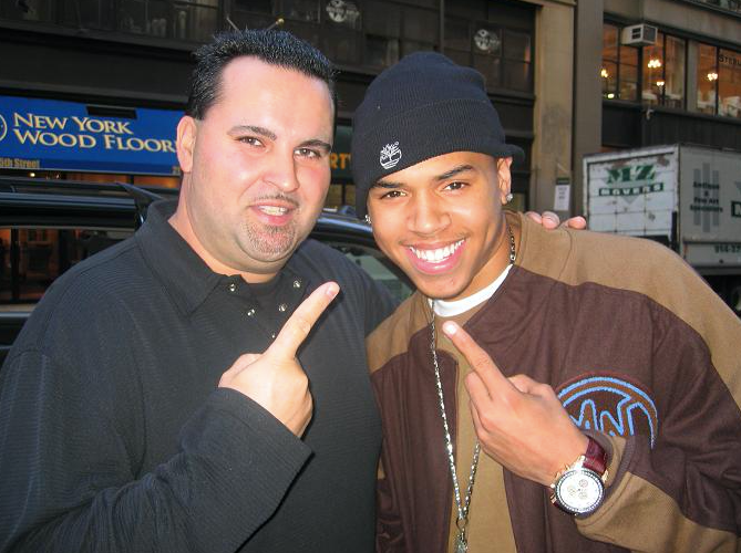 Man and Chris Brown smiling, both pointing. Brown wears a beanie and a brown and beige jacket. Outside building.