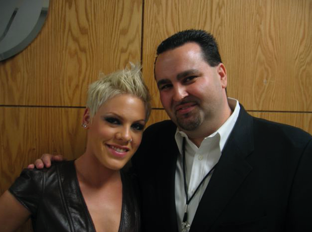 Pink with short blonde hair and a man, posing for a photo against wood paneling.