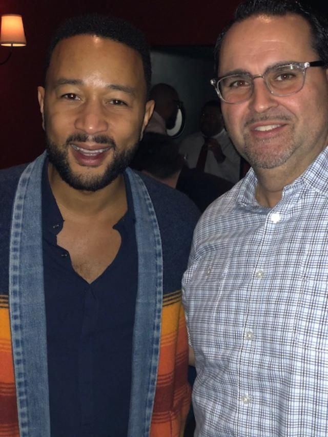 John Legend smiles with a man in glasses; both are indoors, the background blurred.