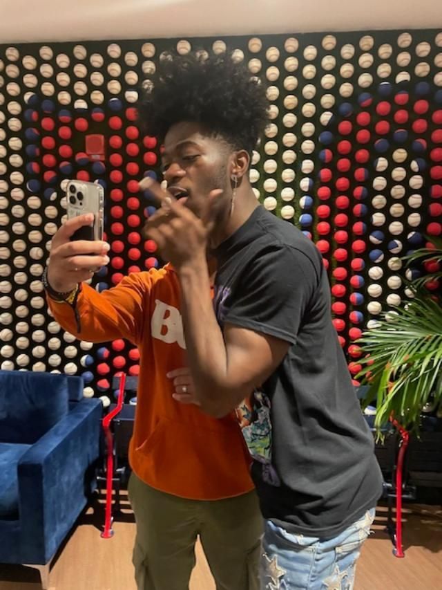 Two people taking a selfie in front of a patterned wall. One wears orange and the other, black.