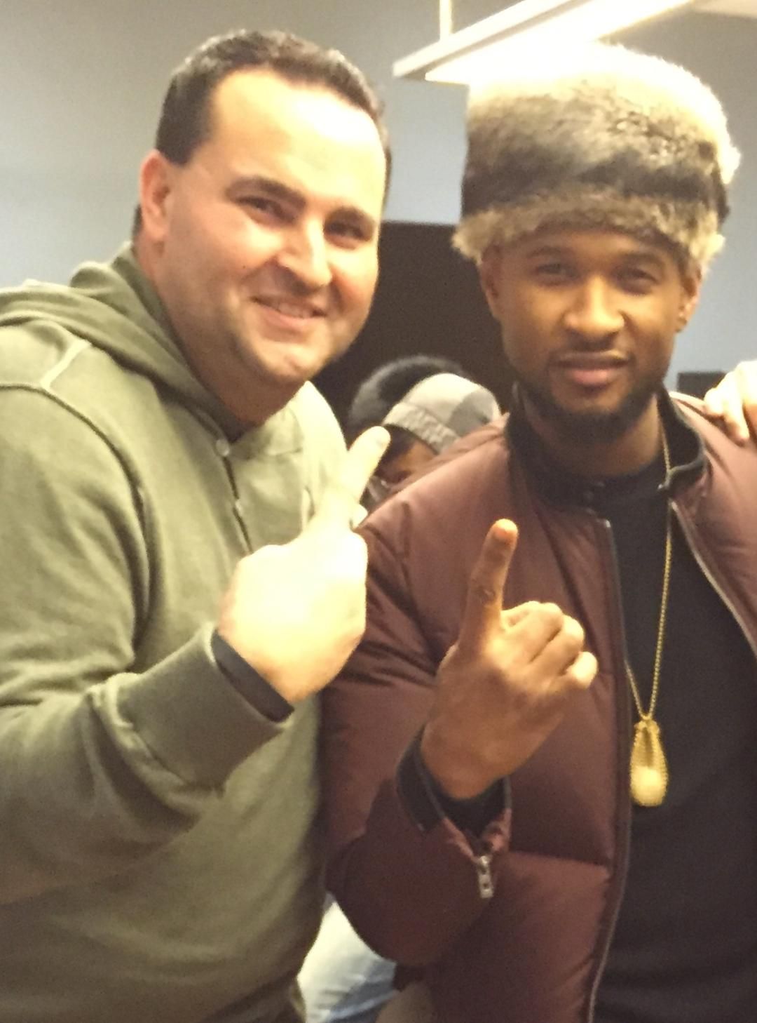 Man in green hoodie and Usher pose together, both holding up one finger.