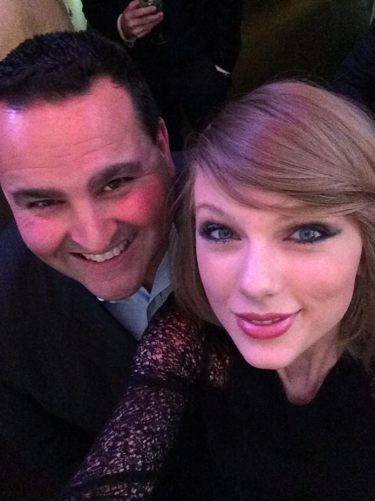 Taylor Swift takes a selfie with a smiling man. Both are close up, in a dimly lit setting.