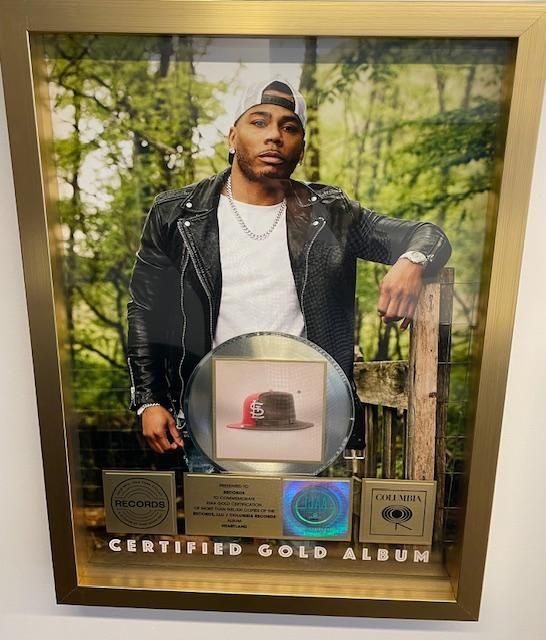 Nelly poses with a framed gold album award. A photo of him is the background. The album is 