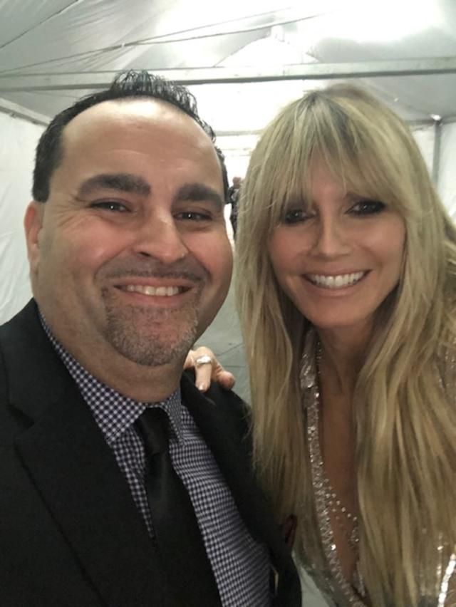 Man smiles next to Heidi Klum, both posing for selfie. They are indoors.