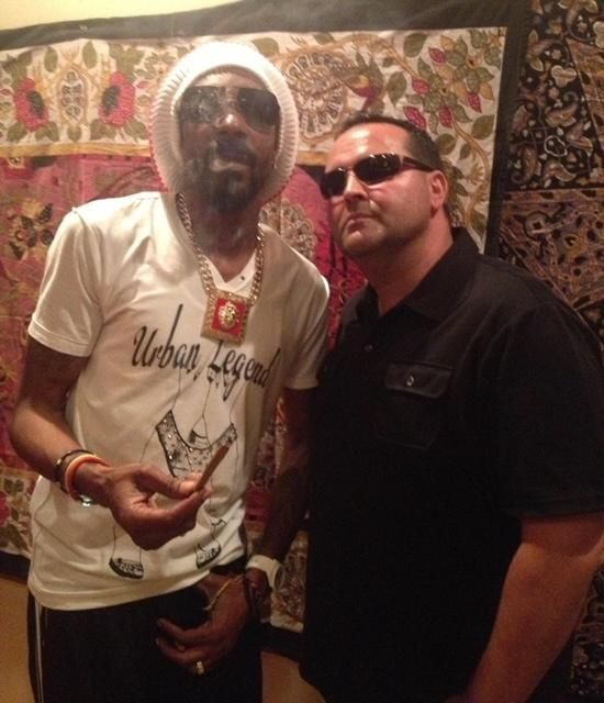 Snoop Dogg and a man in sunglasses pose indoors. Snoop holds a lit object, wearing a white shirt and hat.