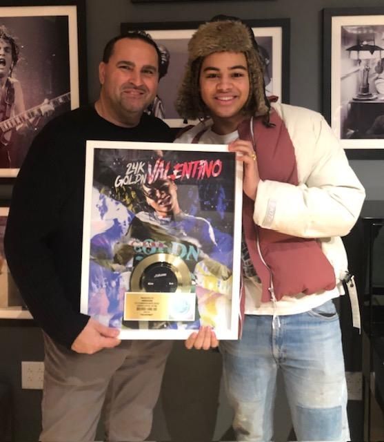 Two people holding a framed gold record in a recording studio. Man in a fur hat smiles.