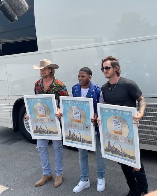 Three men holding gold record plaques in front of a tour bus.