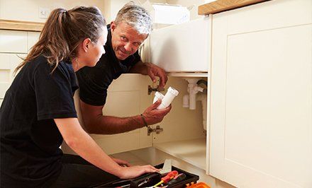 Learn More About Residential Plumbing Two people in a kitchen examine plumbing under a sink, holding white pipes.