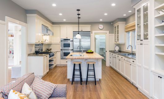 Learn More About Kitchen and Bath Remodeling Spacious cream-colored kitchen with island and bar stools, hardwood floors, and stainless steel appliances.