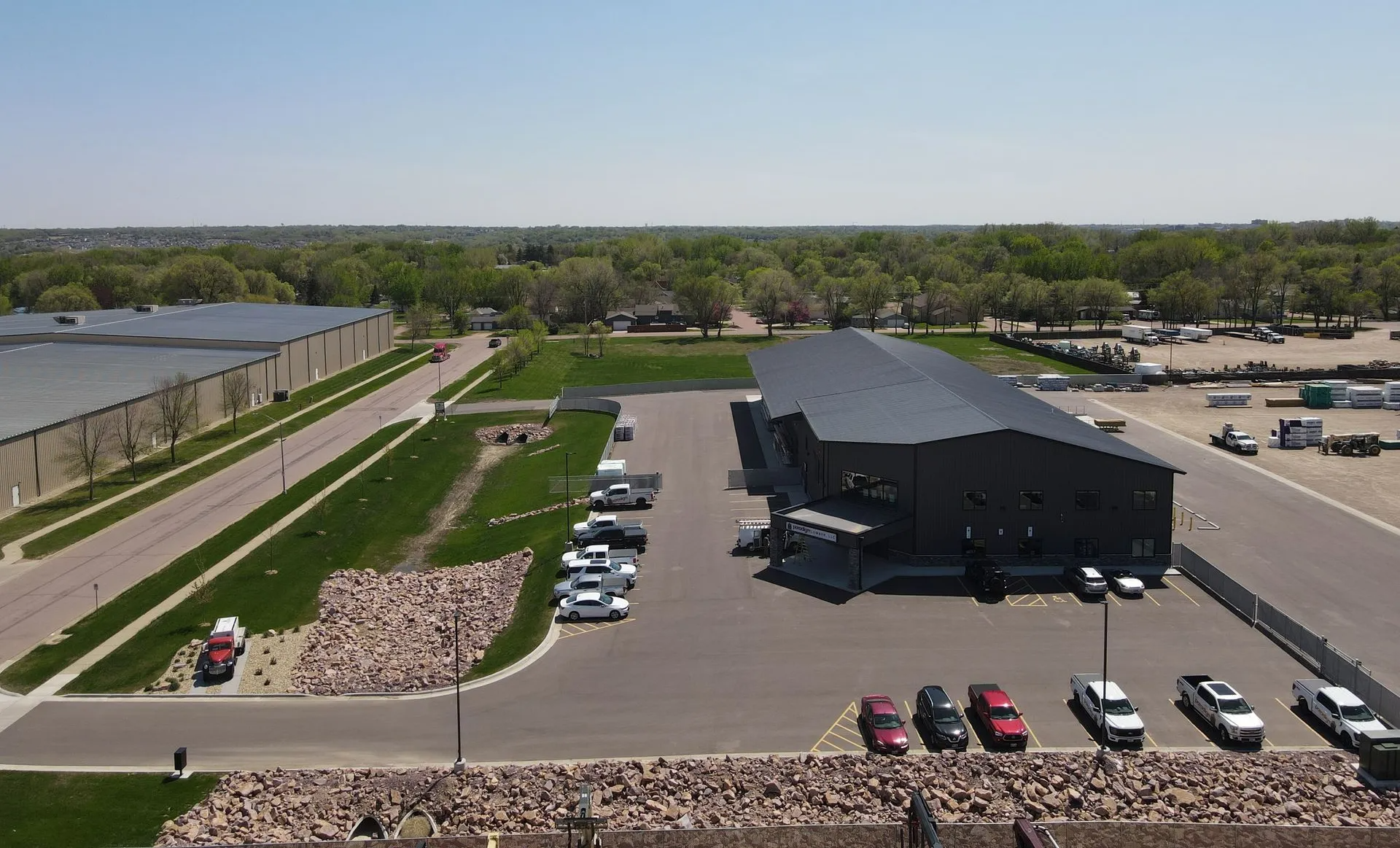 Aerial view of a modern black building with a parking lot in front, trees and clear sky in the background.