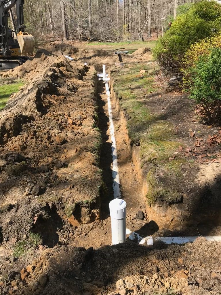 Trench dug beside a road with a white pipe laid in it, next to a tractor and a utility truck.