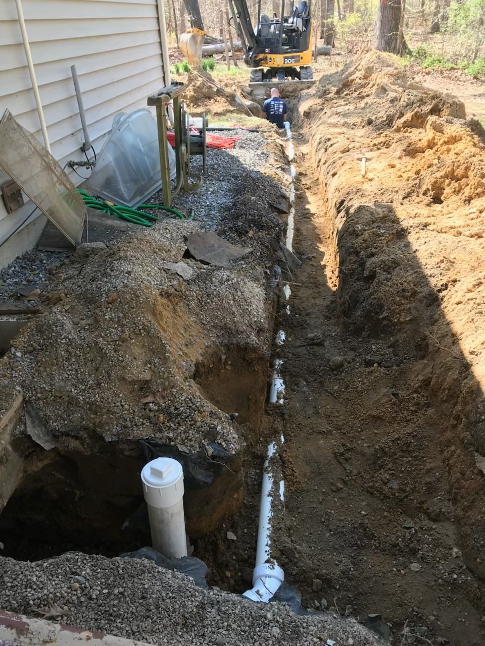 Person standing in a long trench beside a house, with dirt piled along the edge and a truck in the yard.
