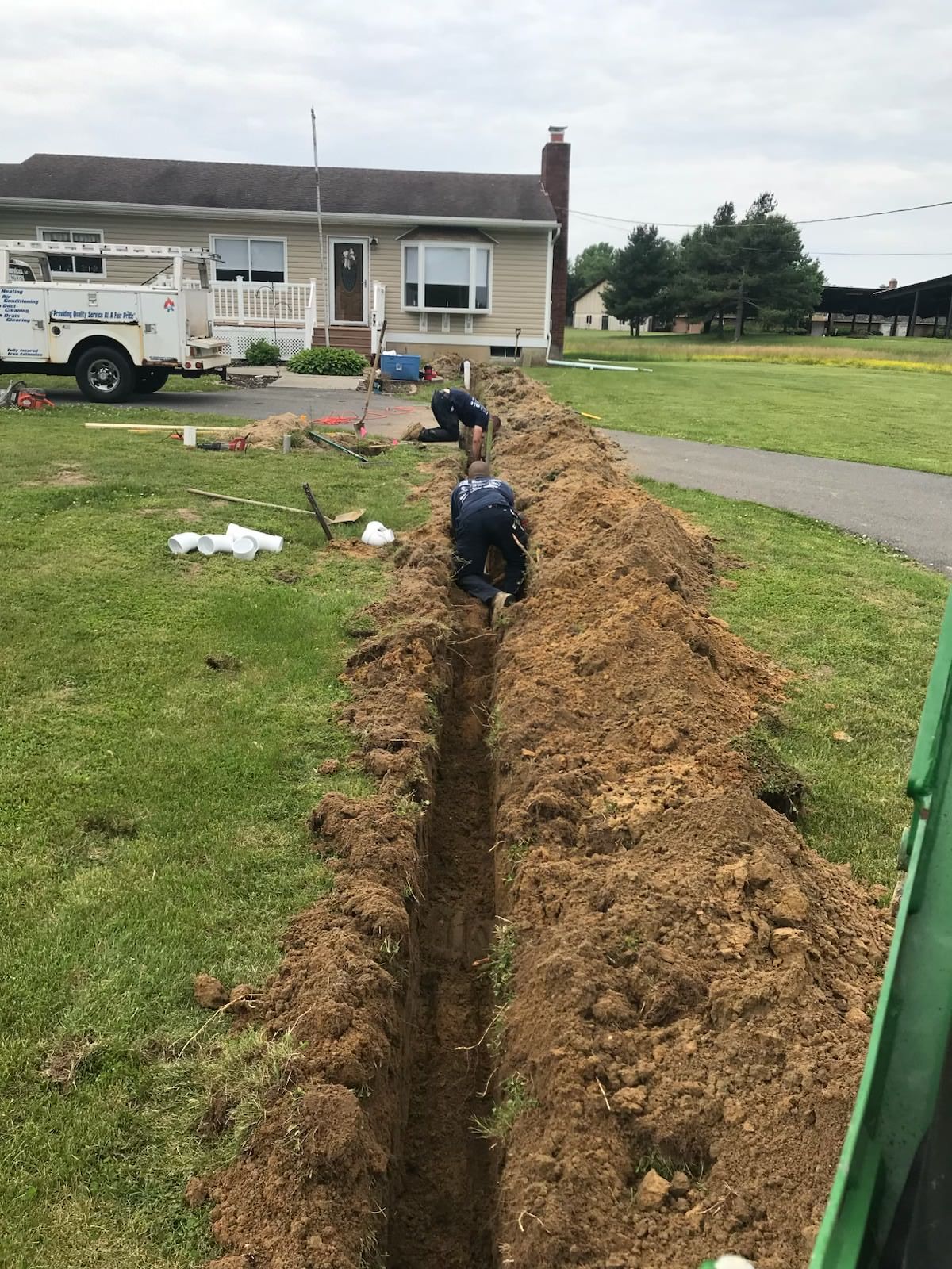 Deep trench beside a house with exposed pipes and excavator work in muddy ground.