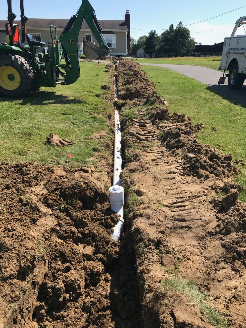 Trench dug along a residential road, with a backhoe on the left and a white truck on the right.