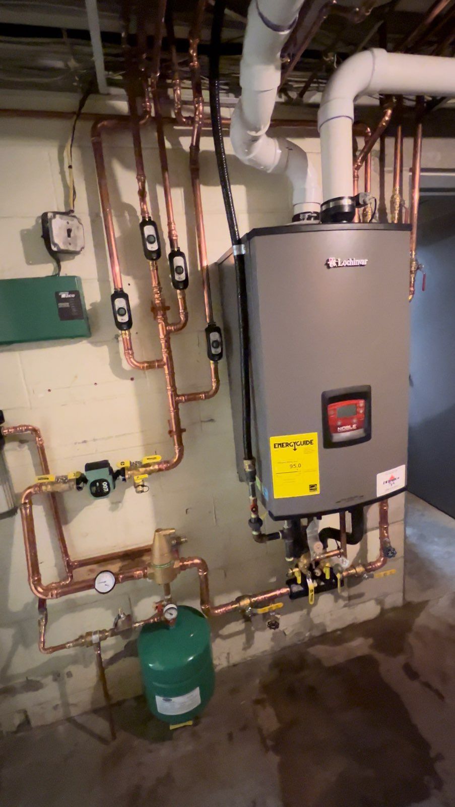 Basement boiler room with wall-mounted furnace, pipes, gauges, and a green tank.