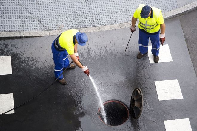 Two workers in yellow vests clear a manhole with a hose on a wet street.