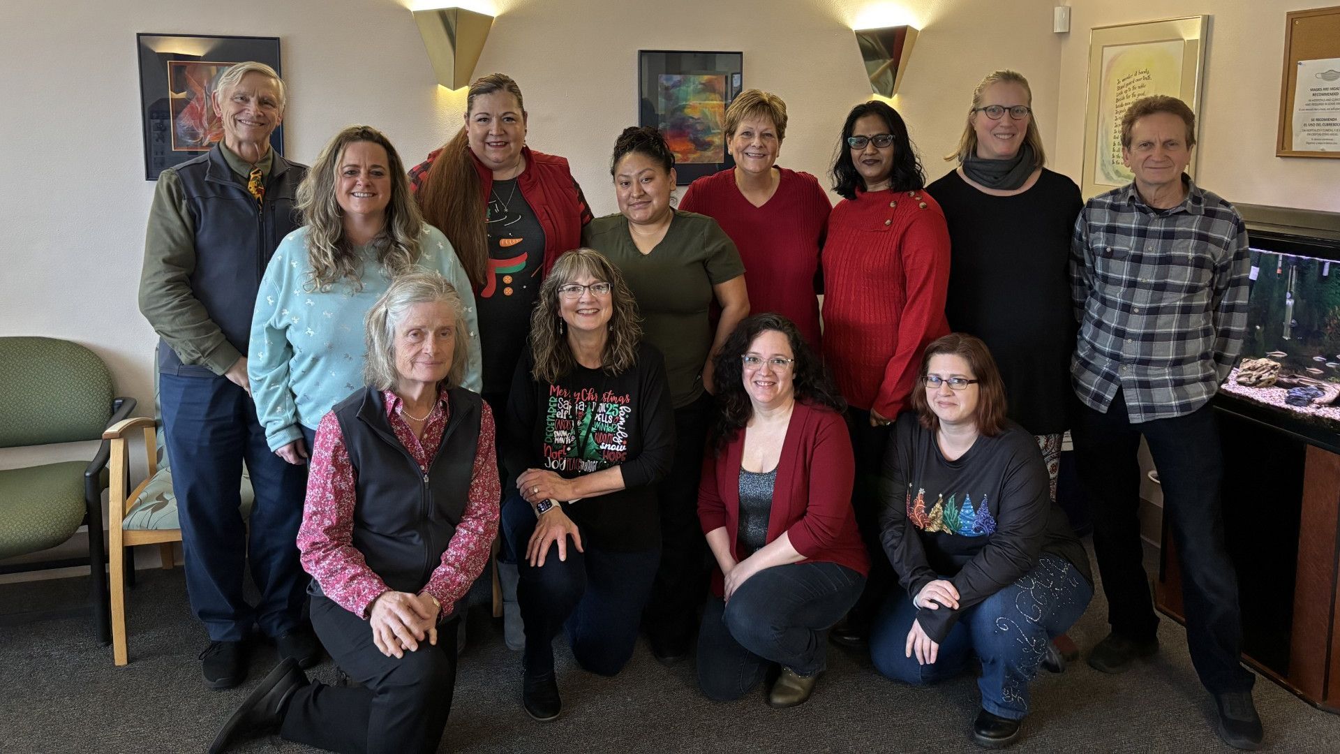 Group of people in a room, some smiling, standing and kneeling. Neutral-toned walls, artwork, and a fish tank are visible.