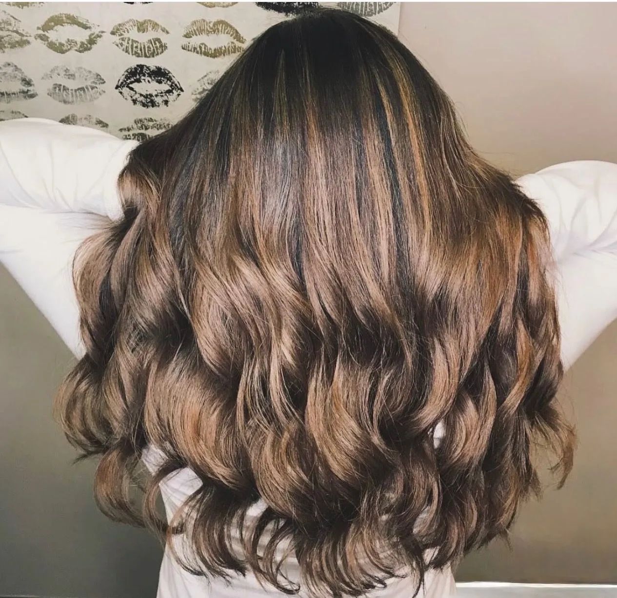 A person seen from behind with long, voluminous, wavy brown hair with highlights against a lip-patterned background.