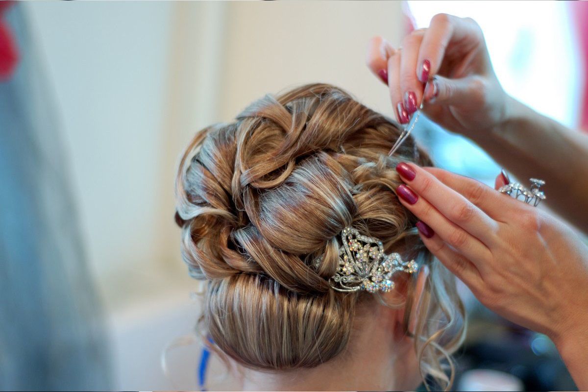 A hairstylist uses a bobby pin to secure an intricate, light brown updo adorned with a decorative, sparkling hairpiece.