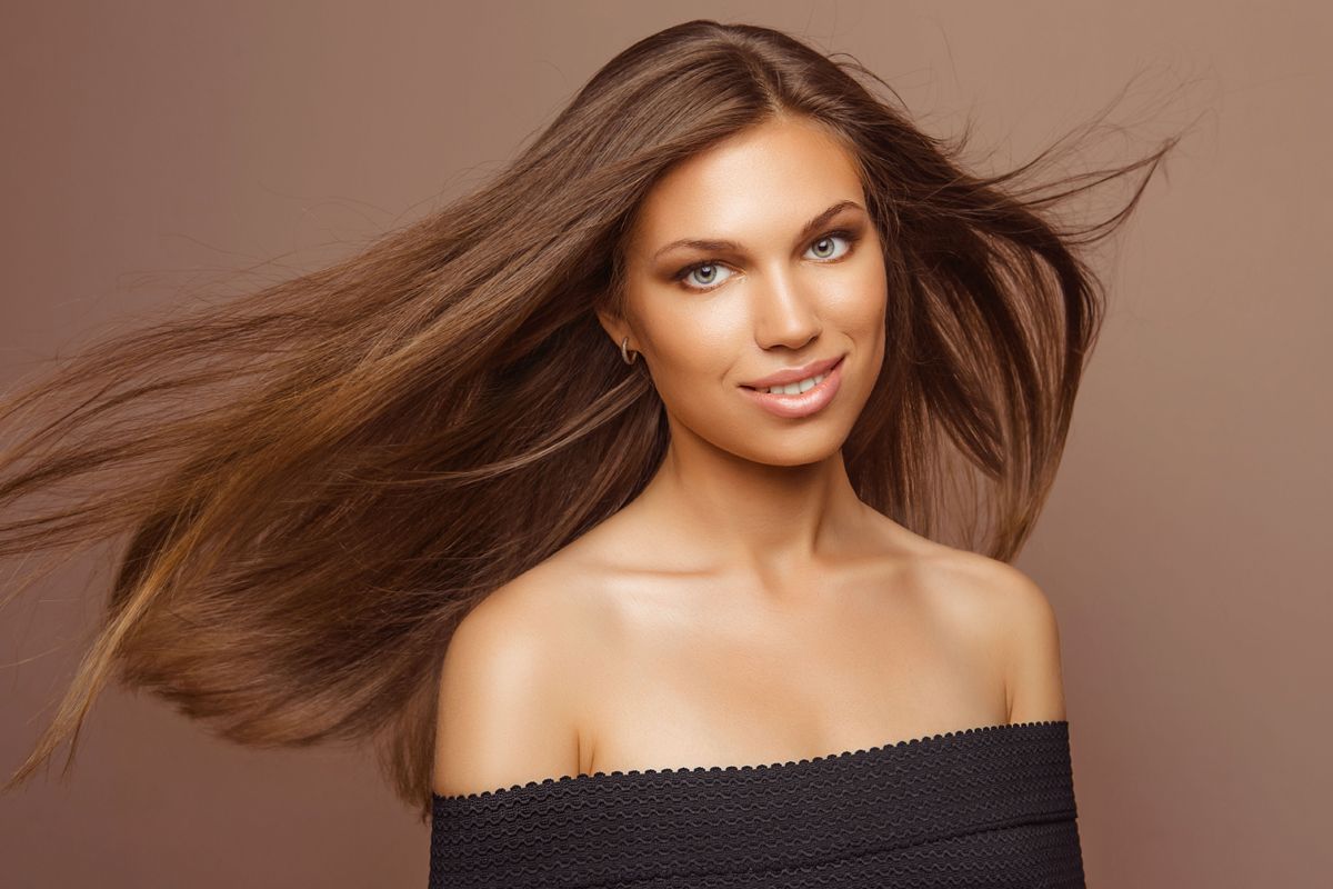 A person with long, flowing brown hair smiles at the camera, wearing a black off-the-shoulder top against a brown background.