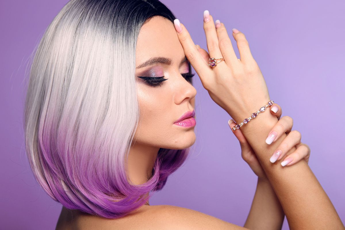 A person with a grey and purple bob hairstyle poses against a purple background, showcasing a bracelet and ring.