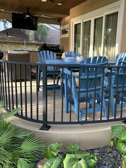 Black railing surrounds a patio with blue chairs and a table. The patio has a TV and faces a yard.