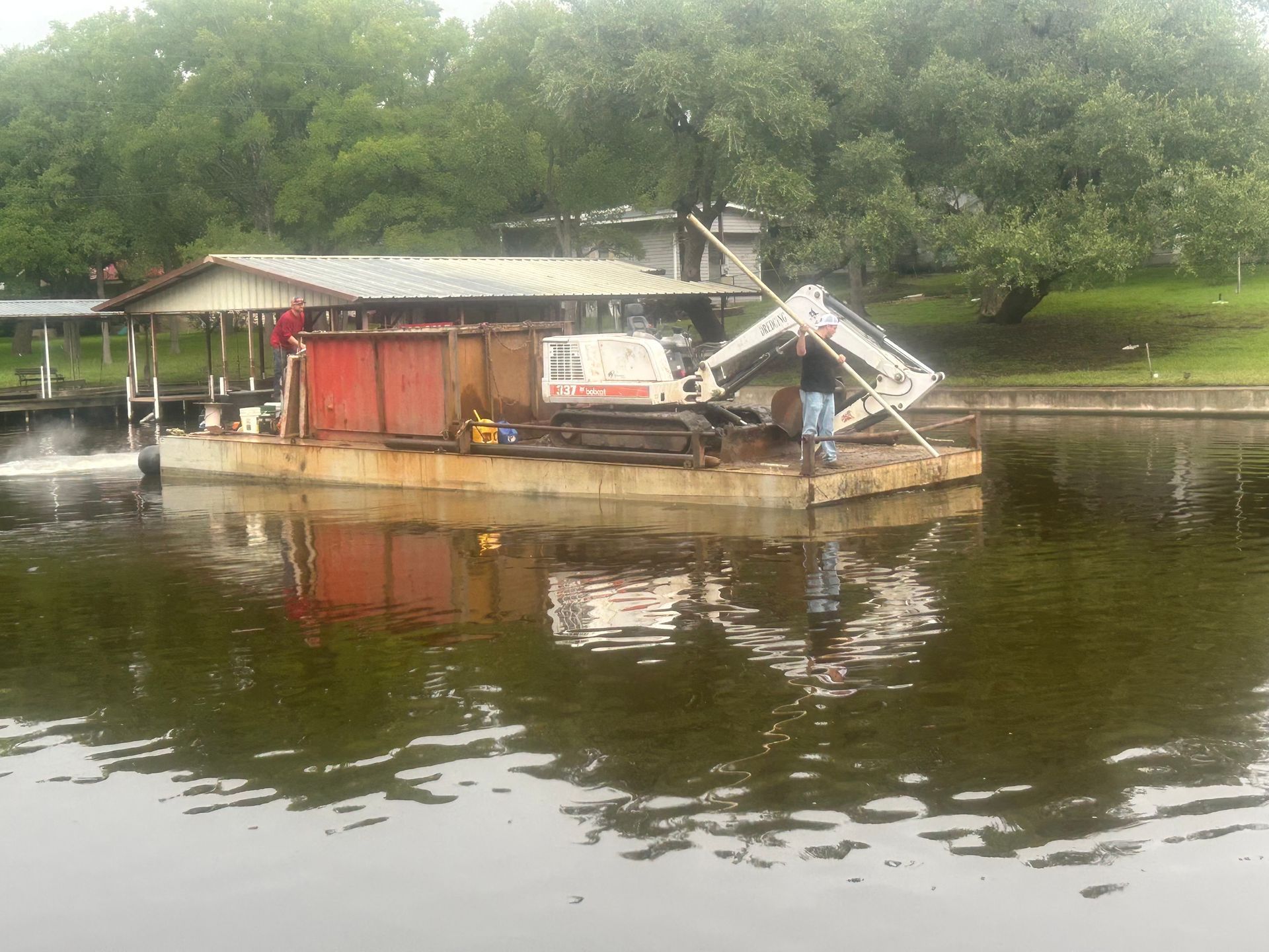 Excavator on a barge in water near a pier, trees in background, working on something.