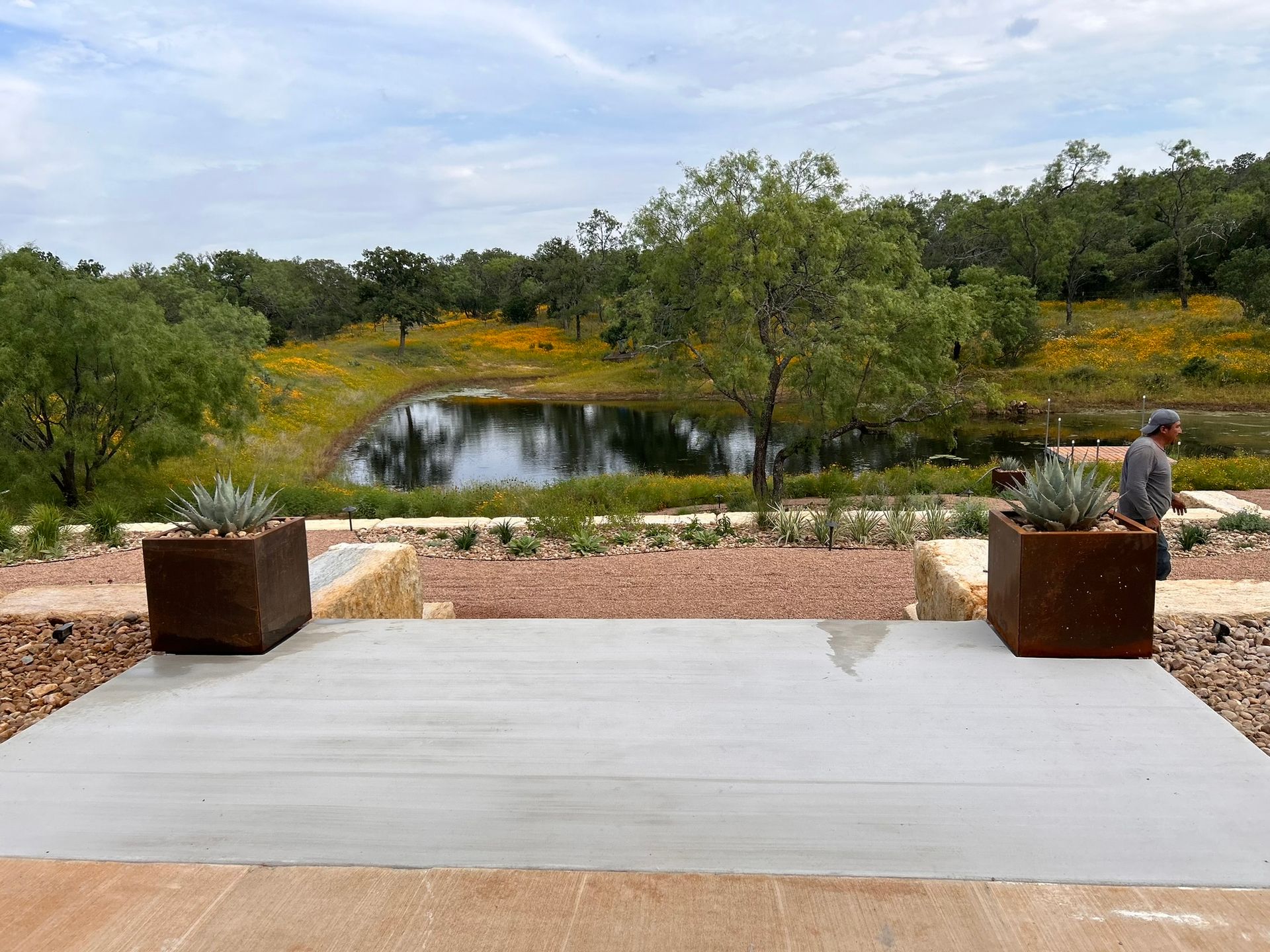 A man is standing in front of a pond with trees in the background