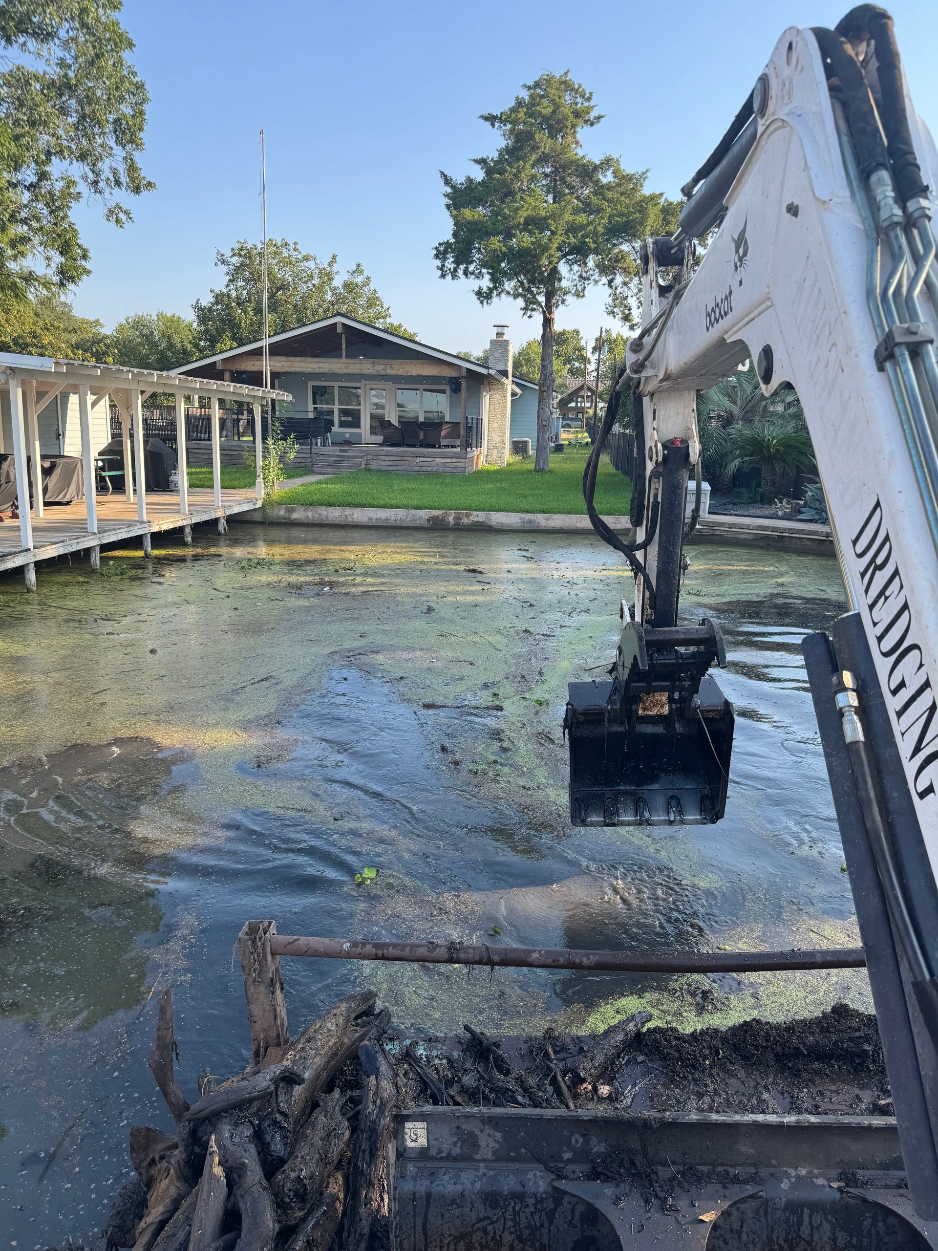 An excavator scoops debris from a murky canal near a house with a dock.