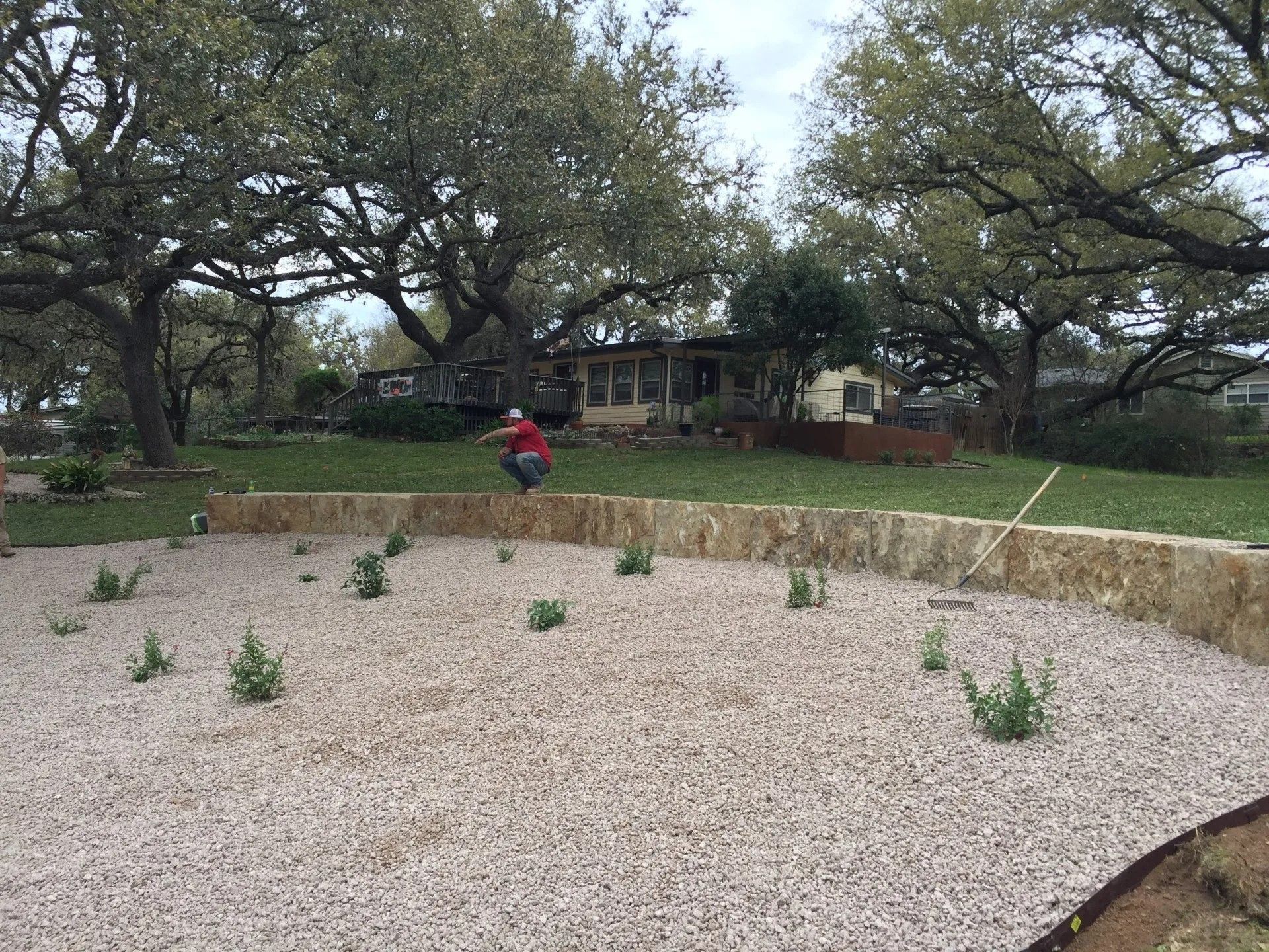 A man is squatting in a gravel area in front of a house.
