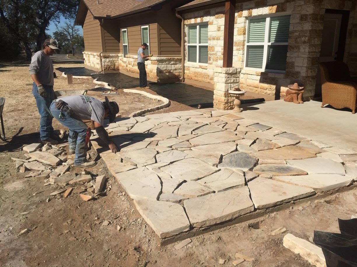 Two men are working on a stone patio in front of a house.