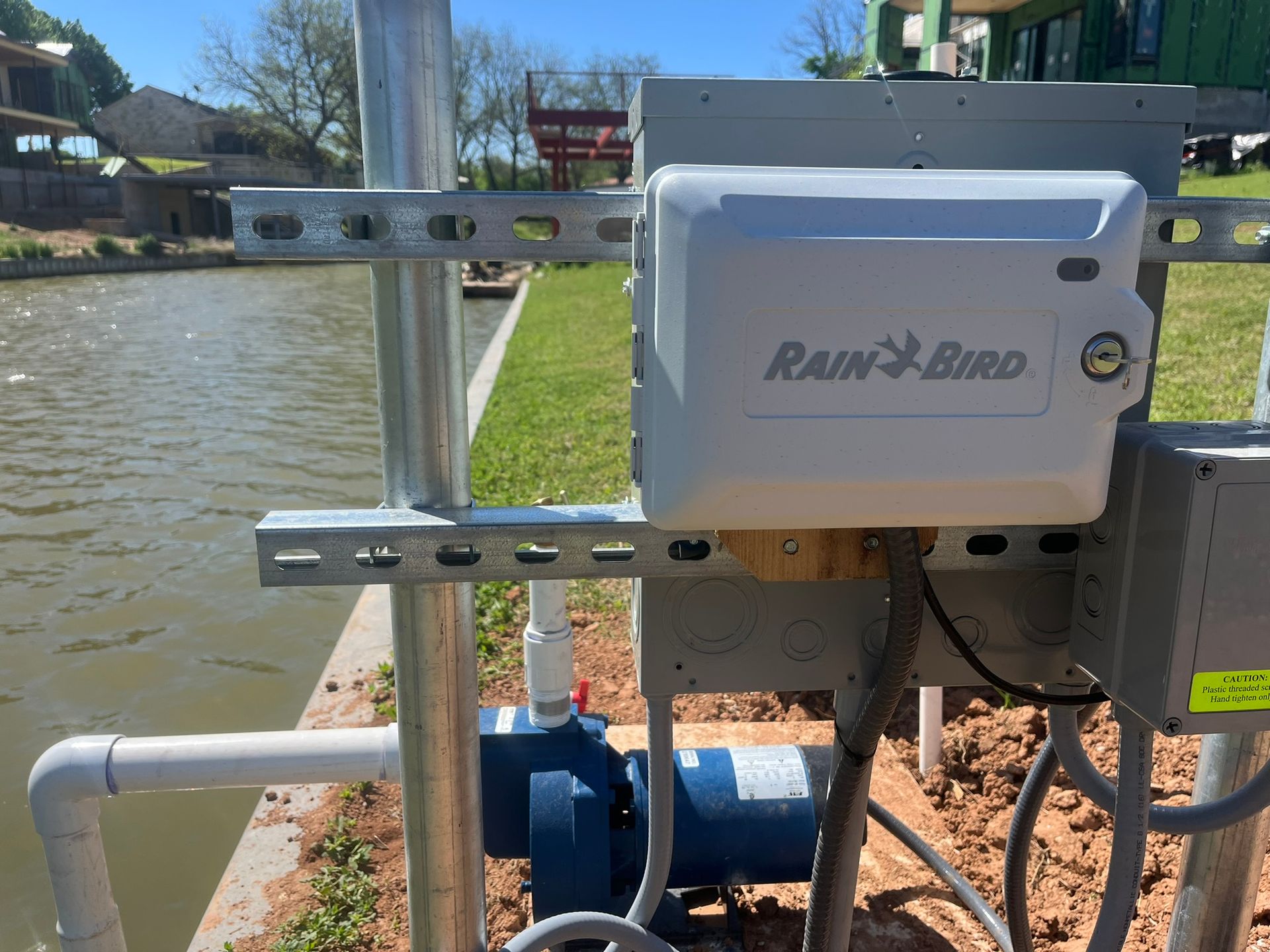 A rain bird box is attached to a metal pole next to a pond.
