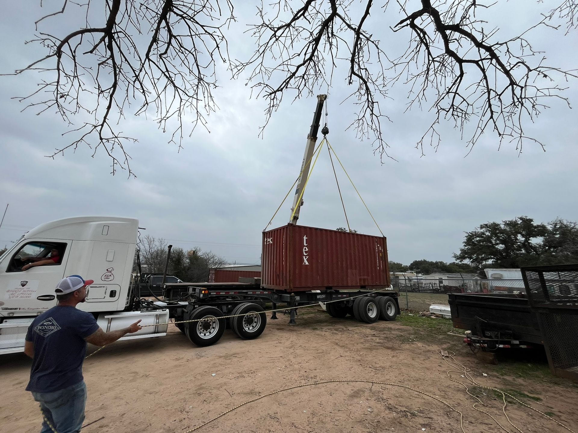 A man is standing in front of a semi truck that is lifting a container