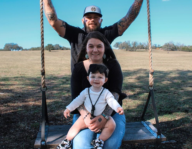Family portrait: Father with tattoos behind a woman holding a child on a wooden swing outdoors under a blue sky.