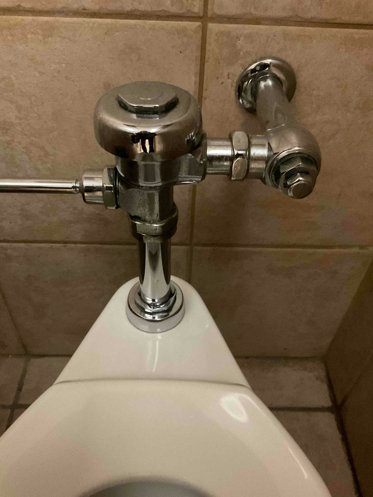 Chrome urinal flush valve mounted on tiled bathroom wall above white urinal
